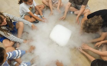 EIFA Eight children sit in a circle on a wooden floor, reaching towards a block of dry ice in the center, surrounded by misty vapor—capturing the excitement and curiosity of International Open Day.