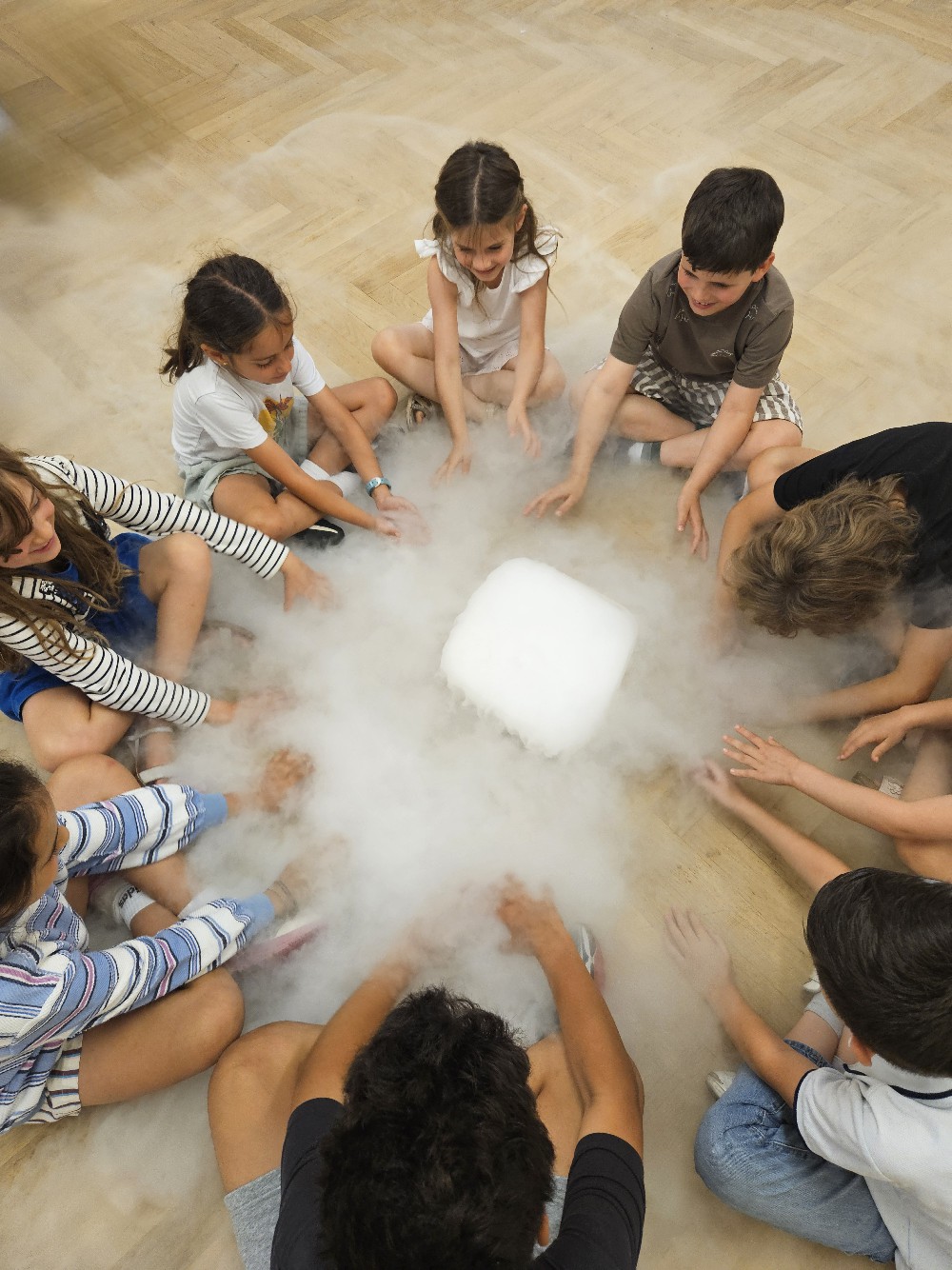 EIFA Eight children sit in a circle on a wooden floor, reaching towards a block of dry ice in the center, surrounded by misty vapor—capturing the excitement and curiosity of International Open Day.