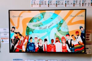 EIFA A classroom display shows diverse children in traditional clothing holding hands below a globe. Above them, cursive text reads “World quiz,” “International Open Day,” and “Samedi 16 Mai.” Alphabet and handwriting charts are on the wall.