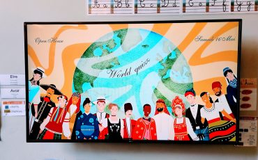 EIFA A classroom display shows diverse children in traditional clothing holding hands below a globe. Above them, cursive text reads “World quiz,” “International Open Day,” and “Samedi 16 Mai.” Alphabet and handwriting charts are on the wall.