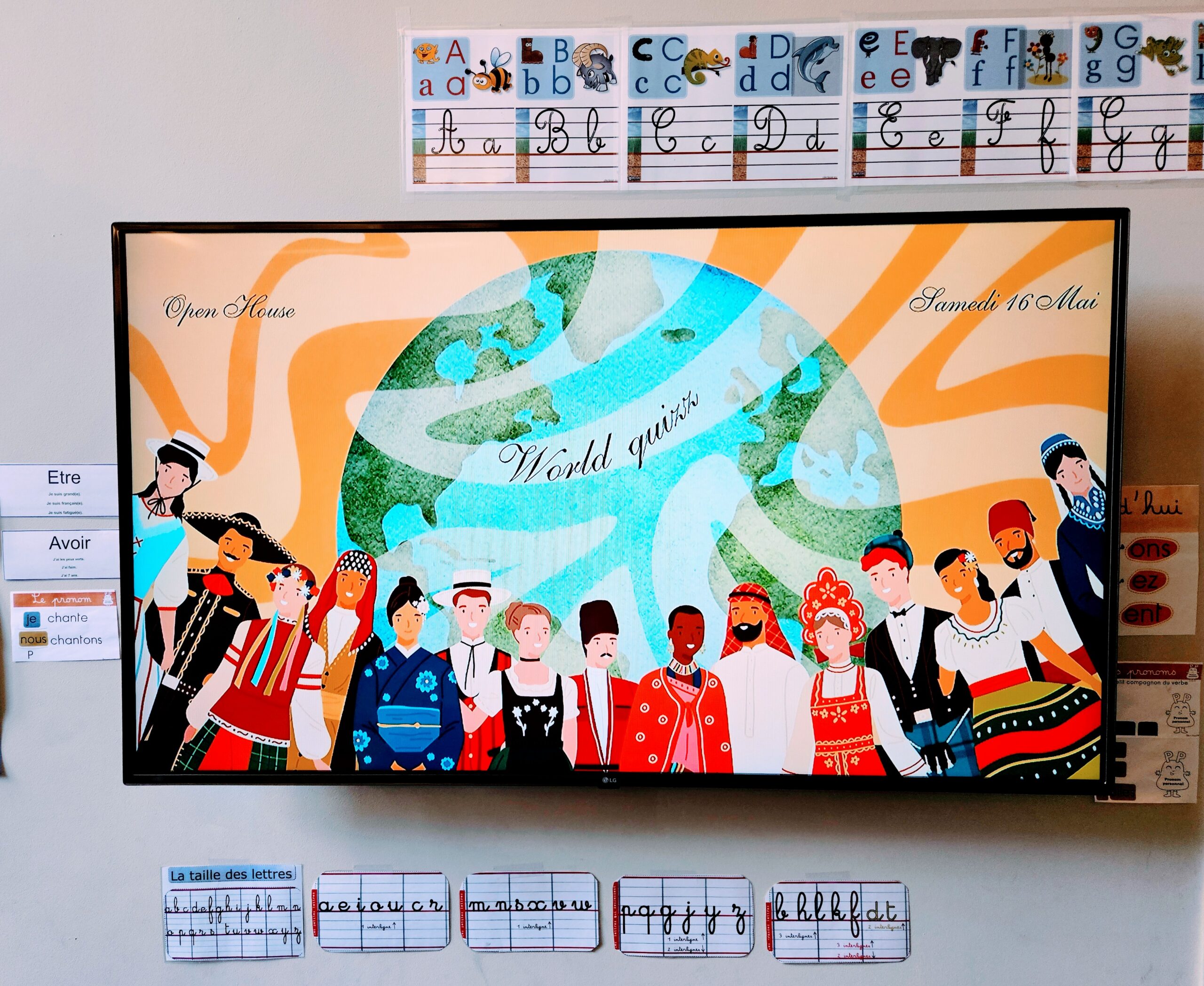 EIFA A classroom display shows diverse children in traditional clothing holding hands below a globe. Above them, cursive text reads “World quiz,” “International Open Day,” and “Samedi 16 Mai.” Alphabet and handwriting charts are on the wall.