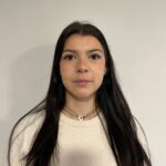 EIFA A young woman with long dark hair and a neutral expression stands in front of a plain light-colored wall, wearing a cream sweater and a silver necklace.