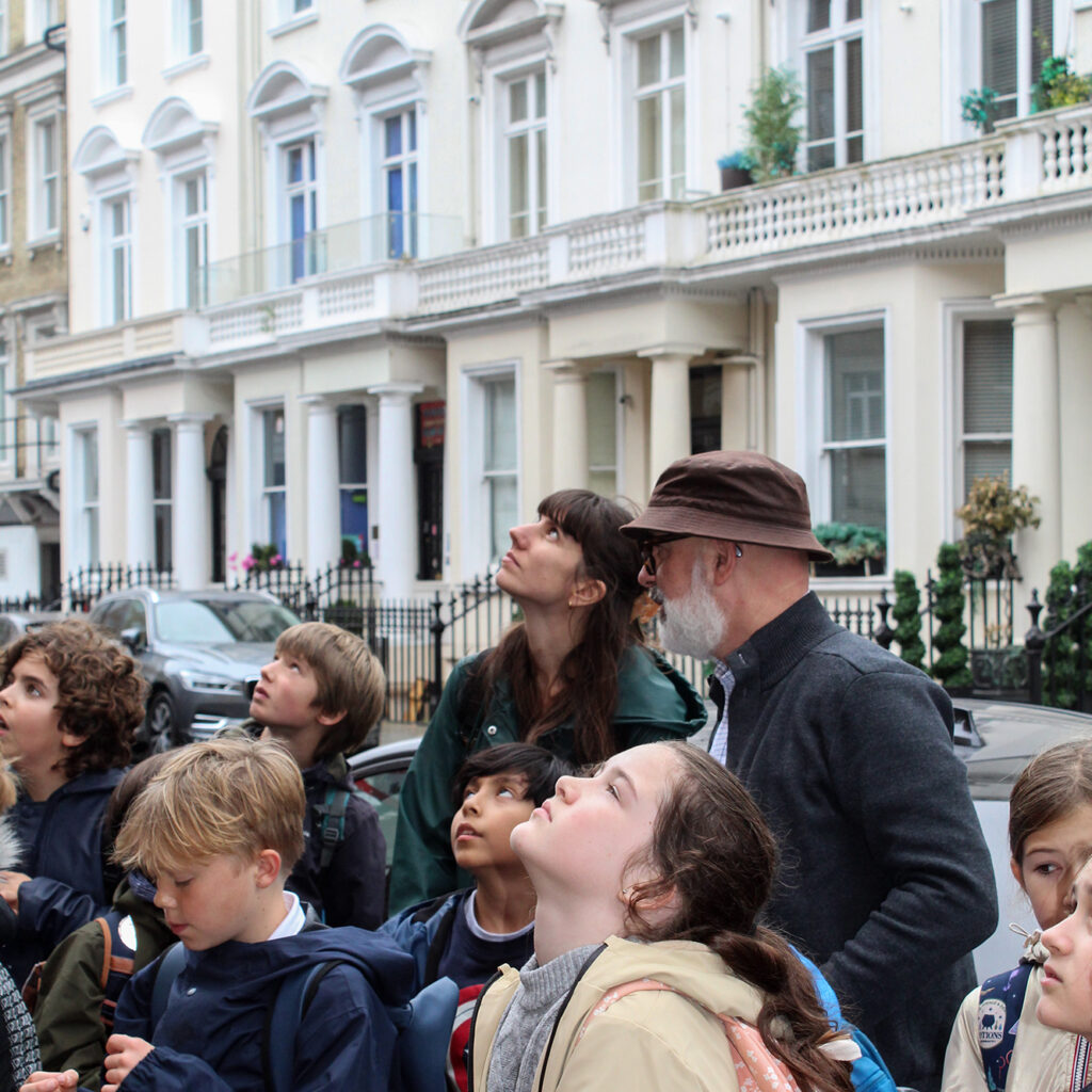 EIFA A group of children and an older man with a beard and hat look up with interest on a city street during International Open Day, surrounded by white terraced houses and parked cars.