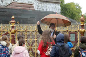 EIFA A group of children with backpacks listens to a man holding an umbrella and gesturing by an ornate fence in front of a large stone monument, taking part in an International Open Day event.