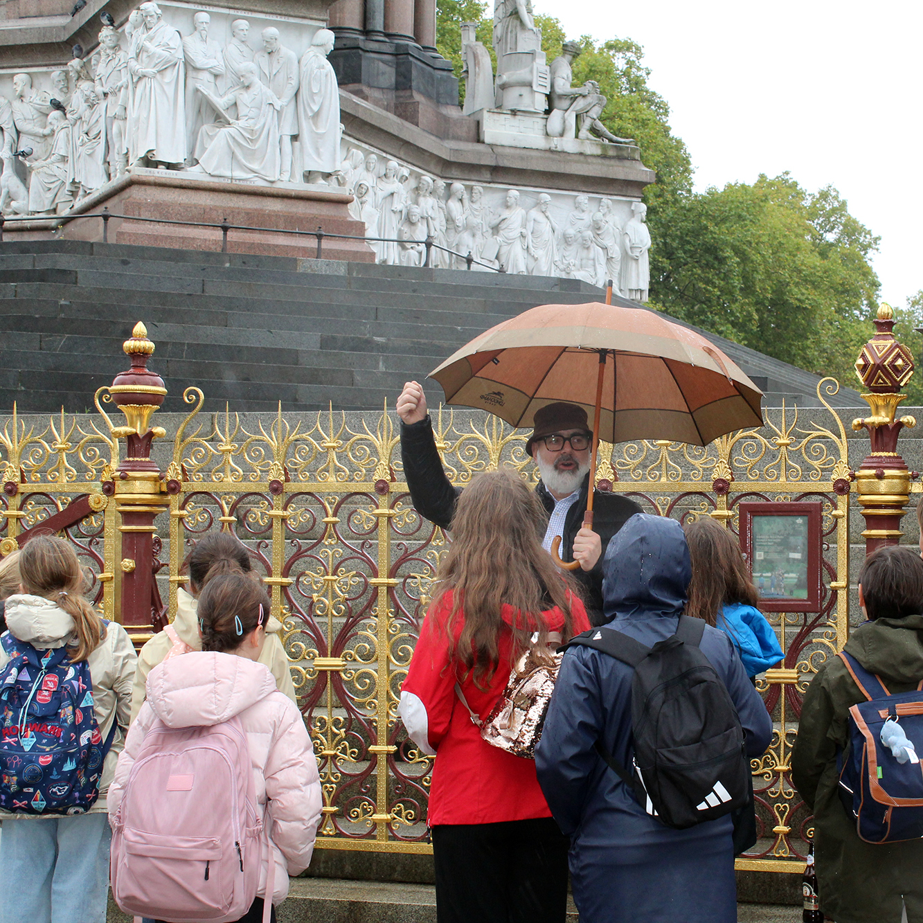 EIFA A group of children with backpacks listens to a man holding an umbrella and gesturing by an ornate fence in front of a large stone monument, taking part in an International Open Day event.