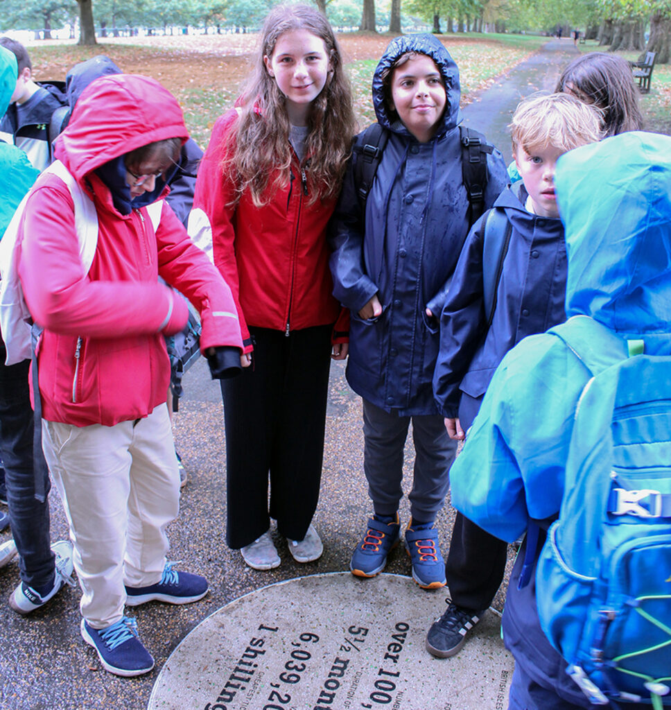 EIFA A group of children in rain jackets stand around a circular plaque set into the walkway in a park during International Open Day. Trees and grass are visible in the background as some children look at the plaque while others face the camera.