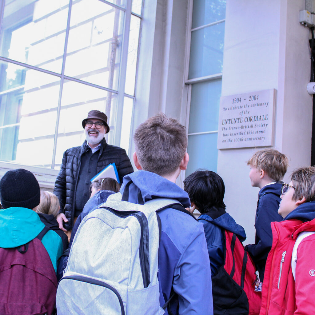EIFA A man wearing a hat and glasses speaks to a group of children with backpacks outside a building, near a commemorative plaque, during the International Open Day.