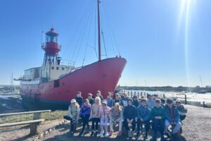 EIFA A group of children sit on a bench in front of a large red ship with a lighthouse tower, docked by the water on a sunny day for International Open Day. The sky is clear and blue.