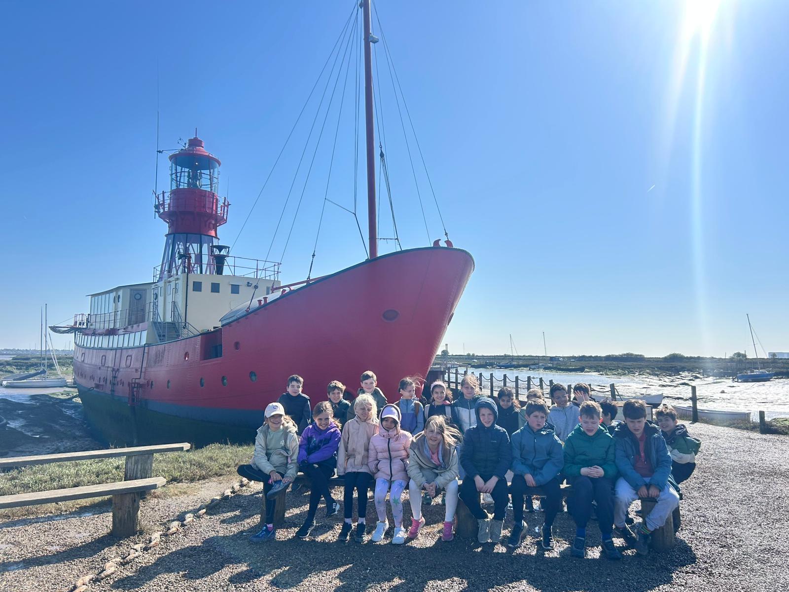 EIFA A group of children sit on a bench in front of a large red ship with a lighthouse tower, docked by the water on a sunny day for International Open Day. The sky is clear and blue.