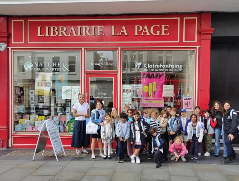 EIFA Un groupe d'enfants et quelques adultes se tiennent debout, tout sourire, devant la Librairie La Page, une librairie française aux murs rouges dont les grandes vitrines exposent des livres et des articles de papeterie.