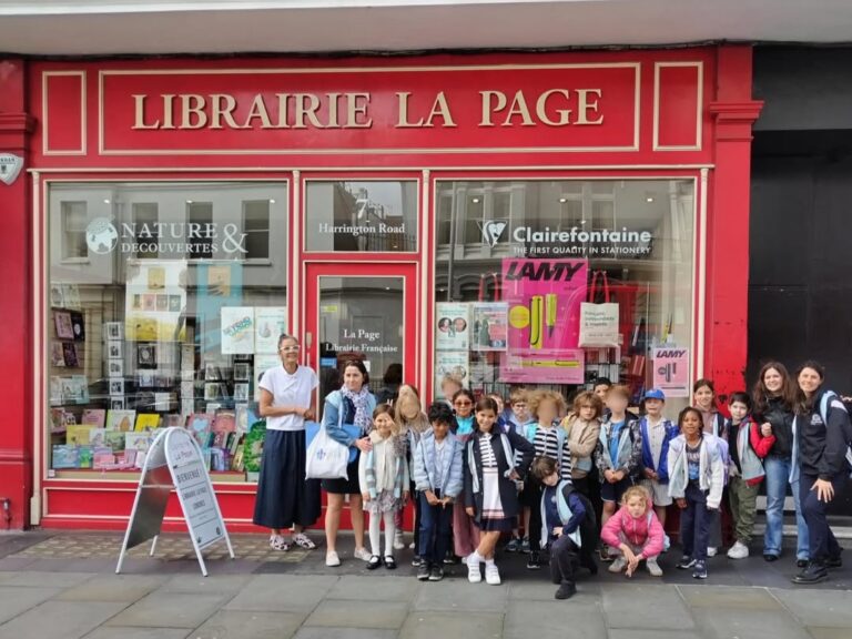 EIFA A group of children and a few adults pose outside the red storefront of Librairie La Page, a French bookstore. The window displays books and stationery, and a sidewalk sign stands near the entrance.