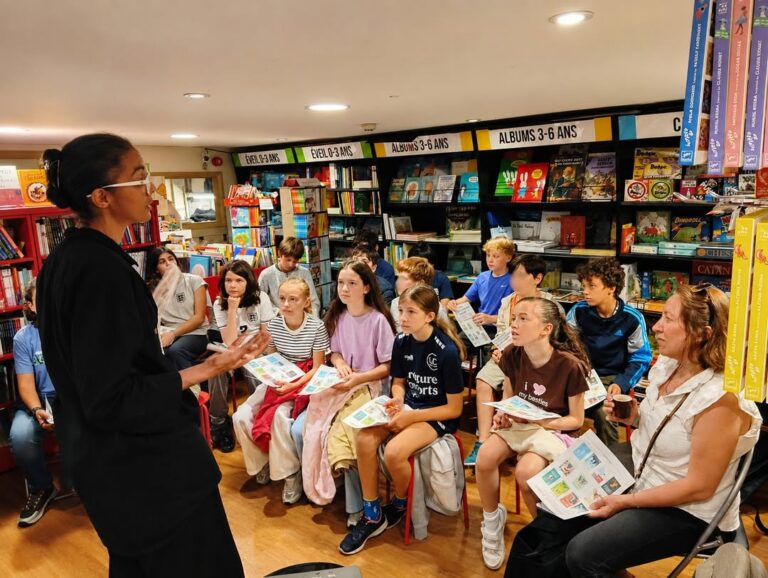EIFA Une femme s'adresse à un groupe d'enfants et d'adolescents assis dans une librairie, chacun tenant des feuilles de papier colorées, avec en arrière-plan des étagères et des couvertures de livres aux couleurs vives.
