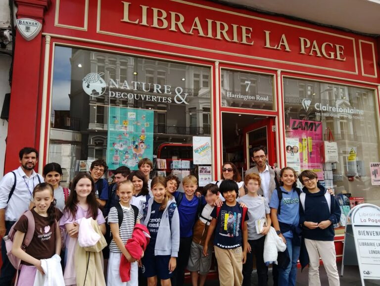 EIFA Un groupe d'enfants souriants et quelques adultes se tiennent devant la devanture peinte en rouge de la Librairie La Page, une librairie française. La vitrine présente des livres et des affiches ; l'ambiance y est animée et conviviale.