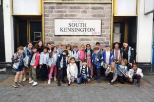 EIFA Un groupe d'enfants et quelques adultes prennent la pose et sourient pour une photo devant un panneau « Welcome to South Kensington SW7 » accroché à un mur de briques. Les membres du groupe sont debout ou à genoux sur un trottoir pavé.