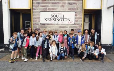 EIFA A group of children and several adults pose and smile in front of a Welcome to South Kensington SW7 sign on a brick wall. The children are dressed casually and stand or kneel together on the pavement.