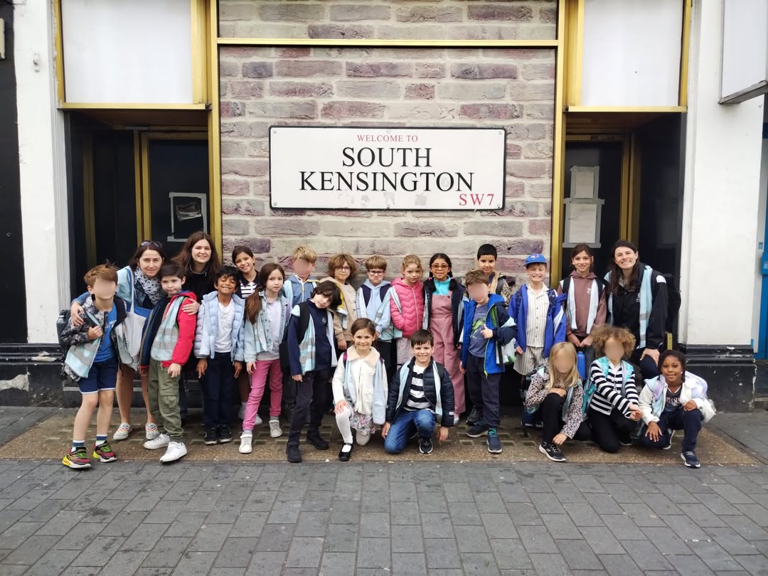 EIFA Un groupe d'enfants et quelques adultes prennent la pose et sourient pour une photo devant un panneau « Welcome to South Kensington SW7 » accroché à un mur de briques. Les membres du groupe sont debout ou à genoux sur un trottoir pavé.
