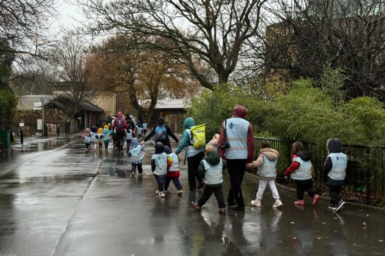 EIFA A group of young children wearing reflective vests walk in a line along a wet path on International Open Day, led by adults also in vests. The rain-soaked ground glistens beneath bare-branched trees lining the route.