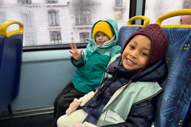 EIFA Two young children in winter clothing smile while sitting on a bus during International Open Day. One wears a teal coat and yellow hat, waving at the camera; the other has a maroon hat and dark coat. The bus window shows a rainy city outside.