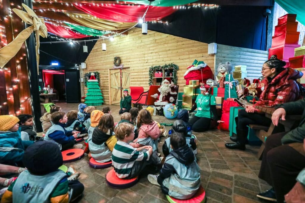 EIFA Les enfants sont assis par terre devant le Père Noël et ses assistants, dans une salle festive décorée de boîtes à cadeaux, de lumières de Noël et de décorations, et écoutent un conte de Noël ou une présentation pour la Journée mondiale de l'alimentation.