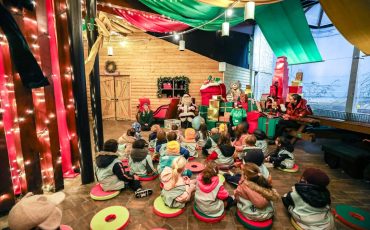 EIFA A group of children sit on colorful cushions, attentively watching performers dressed as elves and Santa in a festive, decorated room with string lights and holiday decorations during International Open Day.