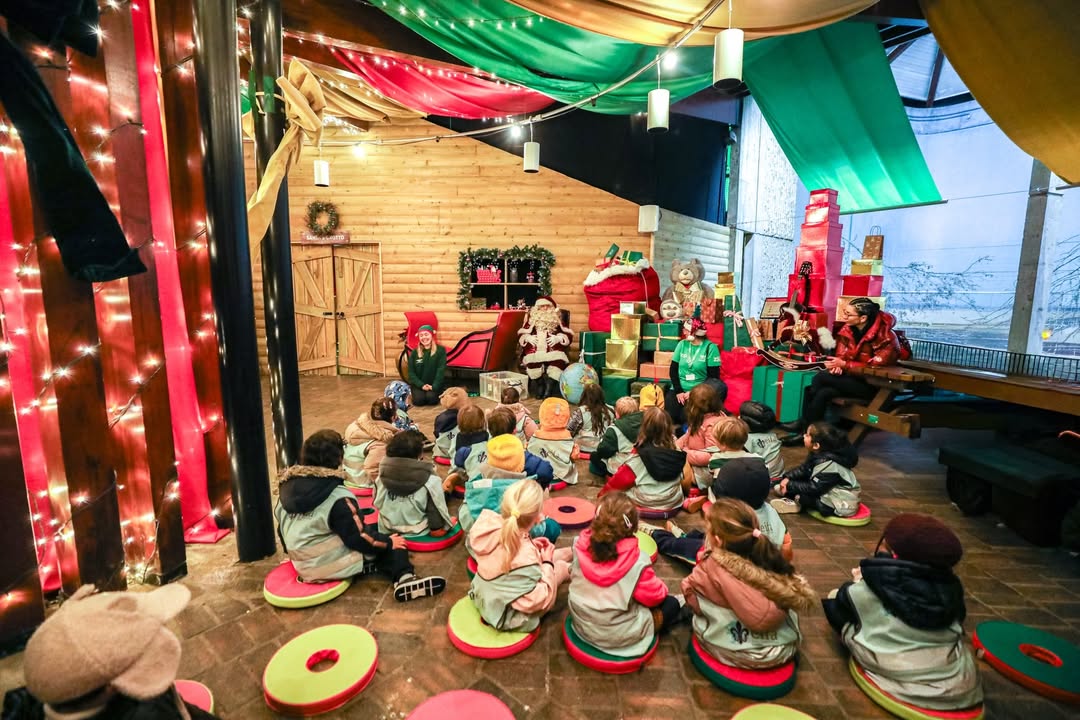 EIFA Un groupe de jeunes enfants est assis sur des coussins colorés, profitant des festivités de la Journée mondiale de l'alimentation. Des artistes costumés en elfes et en Père Noël les ravissent dans un espace intérieur festif avec des lumières, des tissus drapés et des cadeaux emballés.