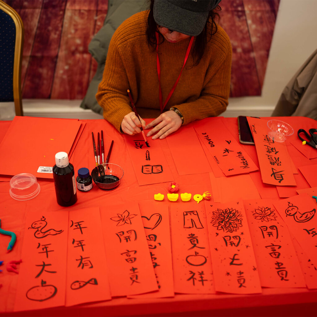 EIFA Une personne est assise à une table rouge et écrit des calligraphies chinoises sur des bandes de papier rouge. La table est remplie doutils de calligraphie, de bouteilles dencre et dobjets sculptés jaunes. La personne porte un pull marron et une casquette.