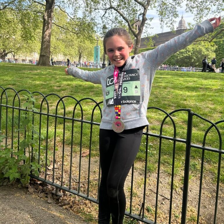 EIFA A smiling young girl stands in a park with her arms outstretched, wearing a race bib and a medal around her neck, celebrating International Open Day. Trees and people are visible in the background behind a black metal fence.