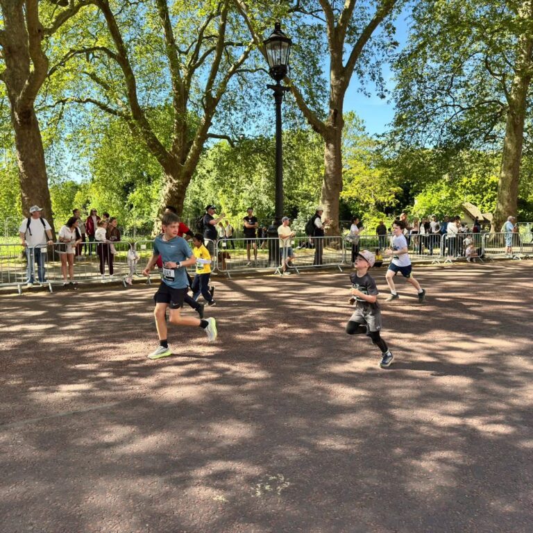 EIFA Children are running a race on a sunlit path lined with trees during International Open Day, while spectators stand behind metal barriers and watch. The park is bright and green, with clear blue skies and dappled shade on the ground.