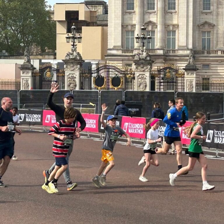 EIFA A group of children and adults are running in a race near Buckingham Palace during International Open Day, with some participants waving and smiling. The palace gates and onlookers can be seen in the background.