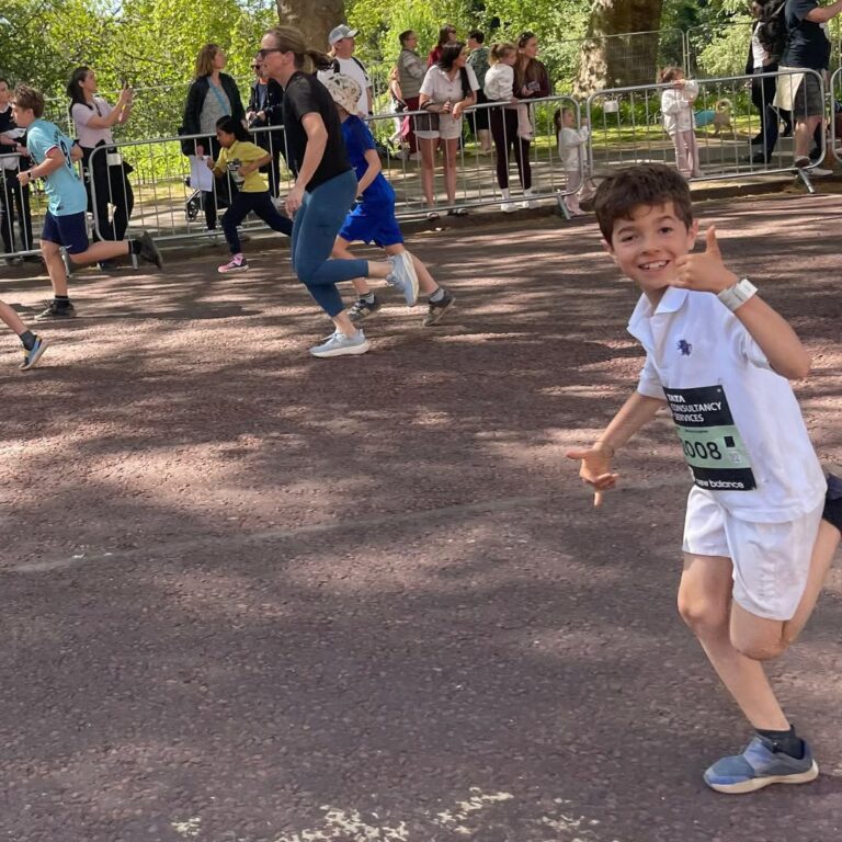 EIFA A cheerful young boy in running gear smiles and gestures playfully at the camera while participating in an International Open Day race, with other runners and spectators in the background on a sunny day.