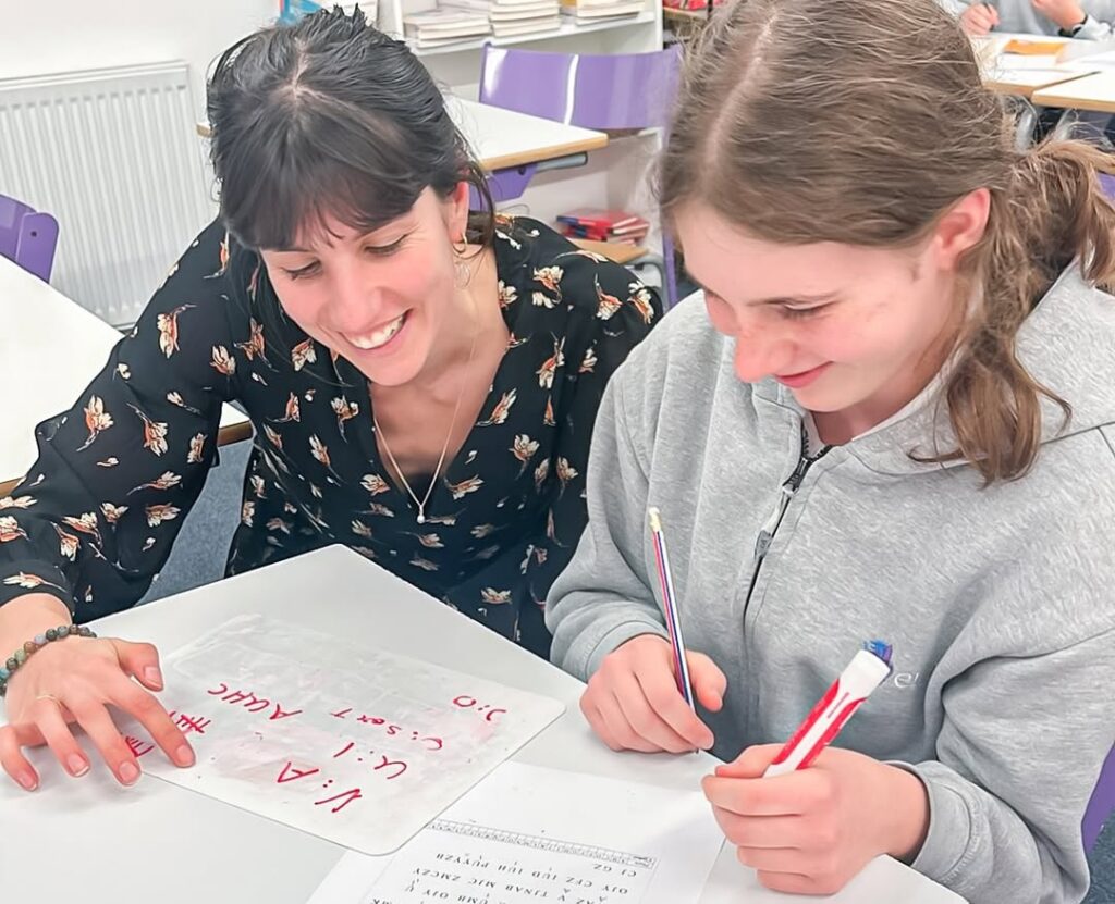 EIFA A teacher and a student sit together at a desk, smiling as they work on an assignment. The student holds a pen and the teacher points to a sheet with handwritten notes. Books and classroom materials are in the background.