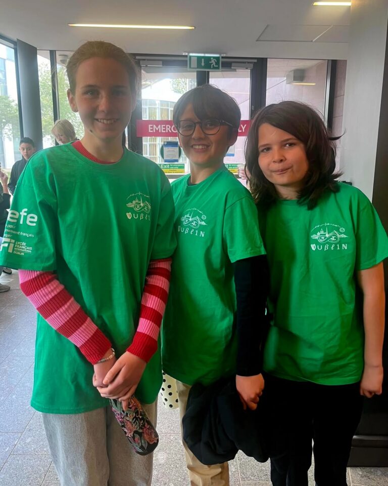 EIFA Three children stand indoors wearing matching green DUBLIN t-shirts, smiling brightly at what appears to be an International Open Day event, with other people and signs visible in the bustling background.