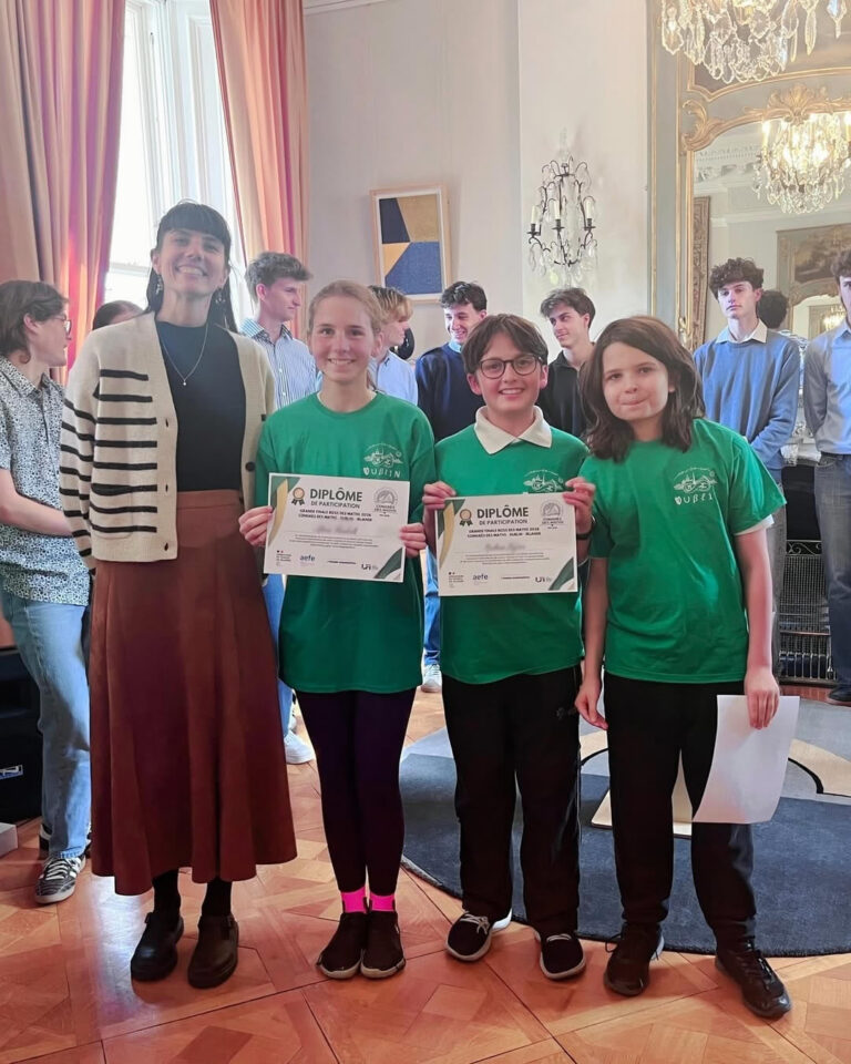 EIFA A woman stands beside three smiling children in green shirts holding certificates, celebrating International Open Day. Several people are in the background of the elegantly decorated room with large windows.