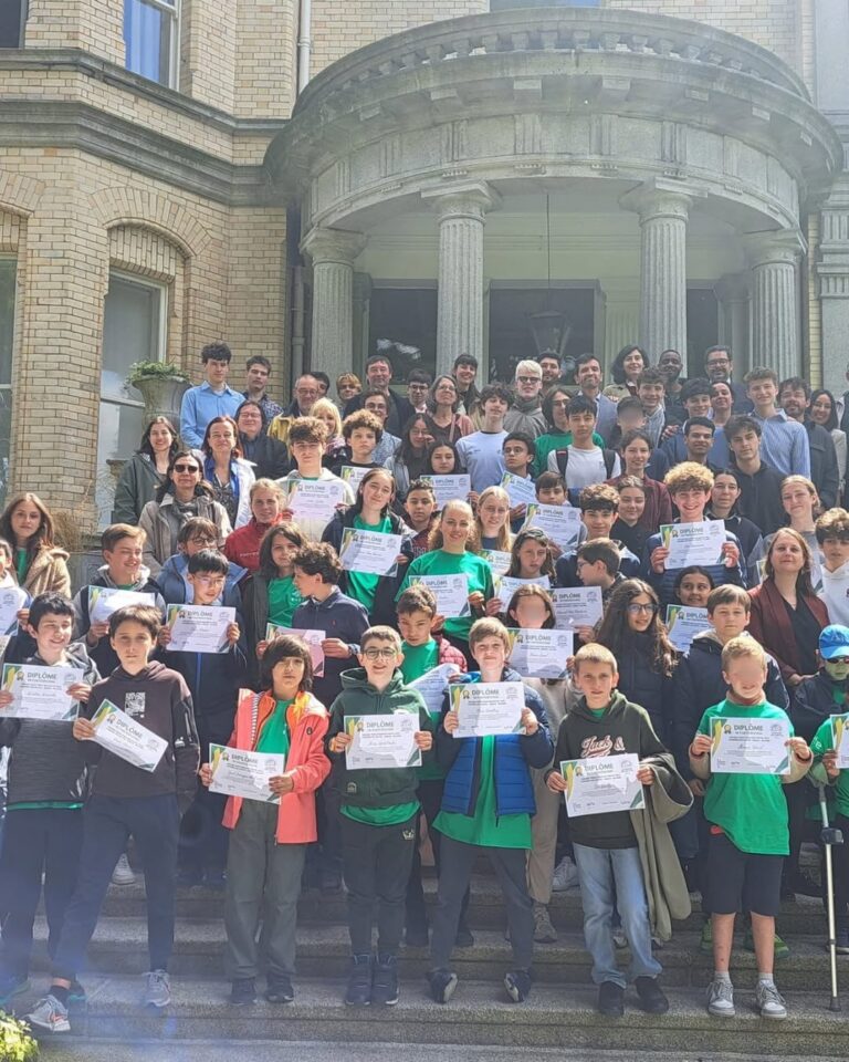EIFA A large group of students and adults stand on steps outside a building, many students in front holding certificates and wearing medals, posing and smiling for a group photo in the sunlight during International Open Day.