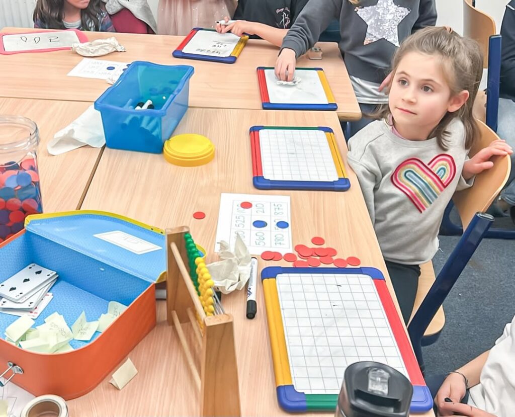 EIFA A group of young children sit around tables, each with a whiteboard, red counters, and other learning tools, appearing engaged in a classroom activity. One girl in a gray shirt with a rainbow heart listens attentively.