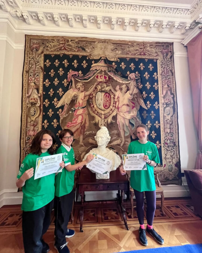 EIFA Three young people in green shirts hold certificates during International Open Day, standing in front of a white bust and an ornate tapestry with a royal crest and fleur-de-lis patterns, in a decorated room with a wooden floor and detailed molding.