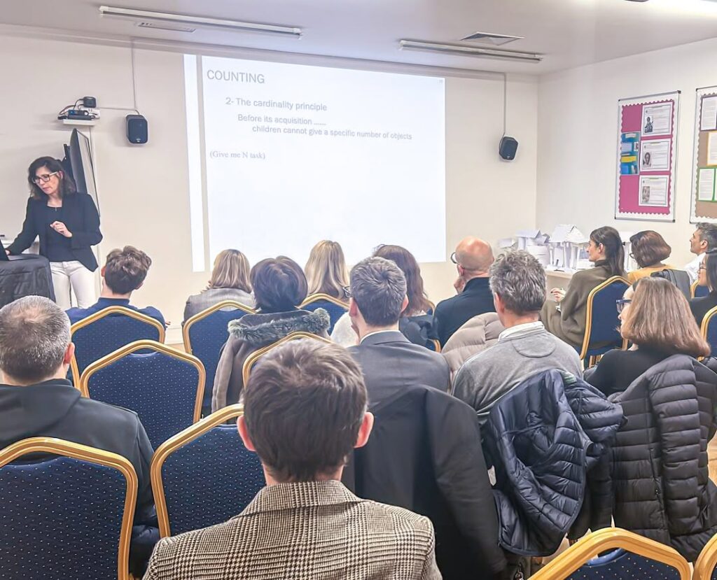 EIFA A woman presents to an audience in a classroom, standing near a projector screen displaying a slide about counting in early childhood. Several adults sit facing the screen, listening attentively.