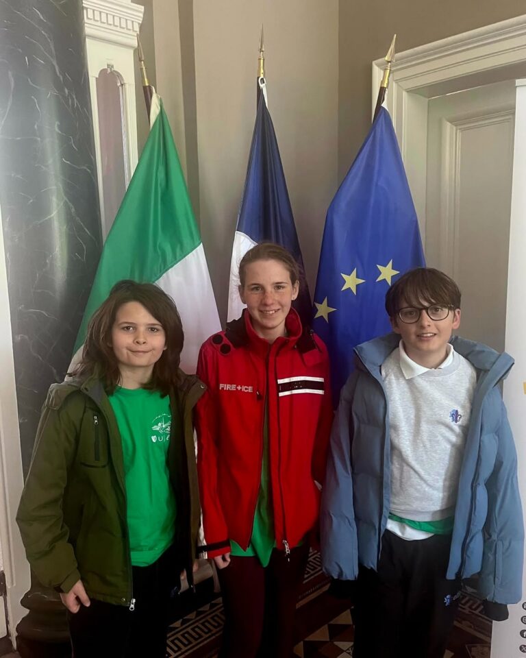 EIFA Three children stand indoors in front of the Italian, French, and European Union flags. Smiling and close together, they're dressed in green shirts with jackets and a red jacketâcelebrating International Open Day.