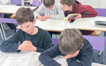 EIFA Four boys sit at desks in a classroom, working together on written assignments. Two boys in the back share a book and paper, while two boys in the front focus on their worksheets. Shelves with books are visible in the background.