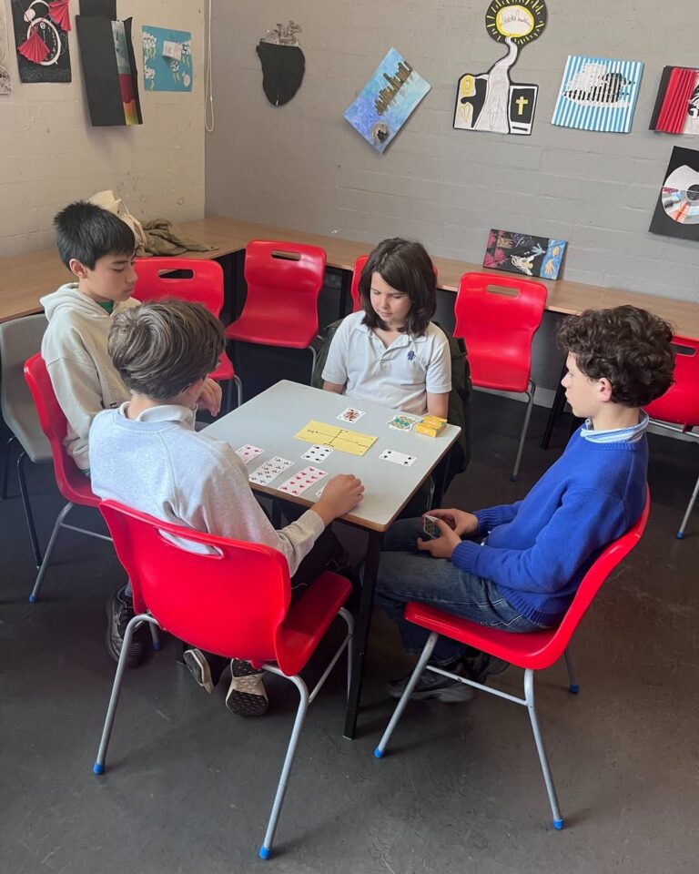 EIFA Four boys sit around a table on red chairs playing a card game in a classroom with artwork on the gray walls and desks in the background, enjoying activities during International Open Day.
