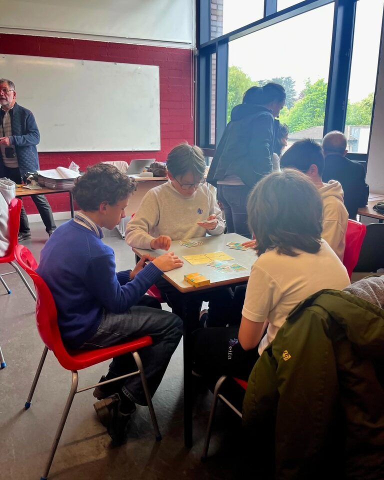 EIFA Four young people sit around a table playing a board game in a brightly lit classroom during International Open Day, while others stand and chat in the background near large windows.