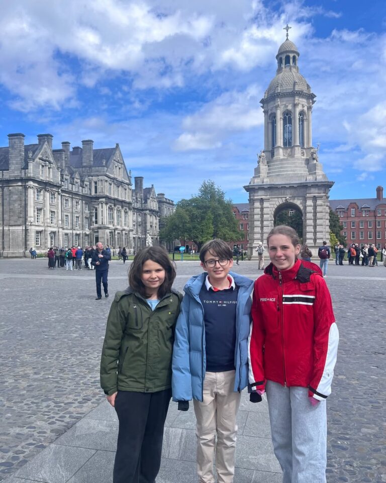 EIFA Three young people stand together on a cobblestone courtyard at Trinity College Dublin during International Open Day, with historic stone buildings and a bell tower in the background, under a partly cloudy blue sky.