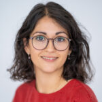 EIFA A person with shoulder-length dark hair and glasses is smiling at the camera in their red top, exuding a warm, bilingual charm. The plain, light-colored background hints at a cozy London studio, perhaps echoing the vibrancy of EIFAs diverse atmosphere.