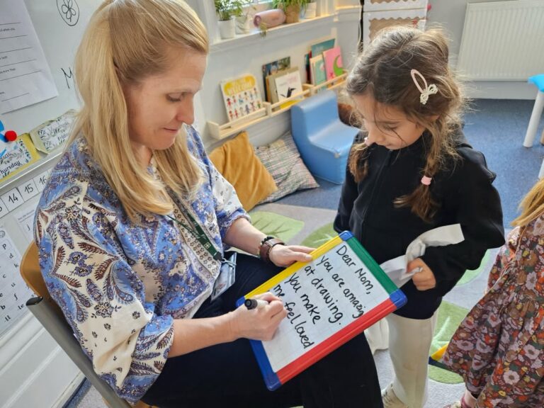 EIFA A woman sits with a young girl, writing a message on a colorful whiteboard that says, Dear Mom, You are amazing. Your creativity makes me feel loved. The girl watches attentively, holding a soft toy.