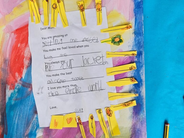 EIFA A child’s handwritten note to their mom is decorated with yellow paper strips and drawings of flowers, placed on a colorful painted background with blue, pink, and red shades. Some crayons are visible in the corner.