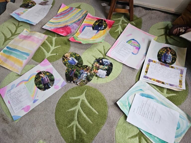 EIFA Children’s artwork and handwritten sheets are spread out on a green leaf-patterned carpet. Some include colorful painted designs, while others have photos of a child and writing attached. A wooden stool is visible at the top.