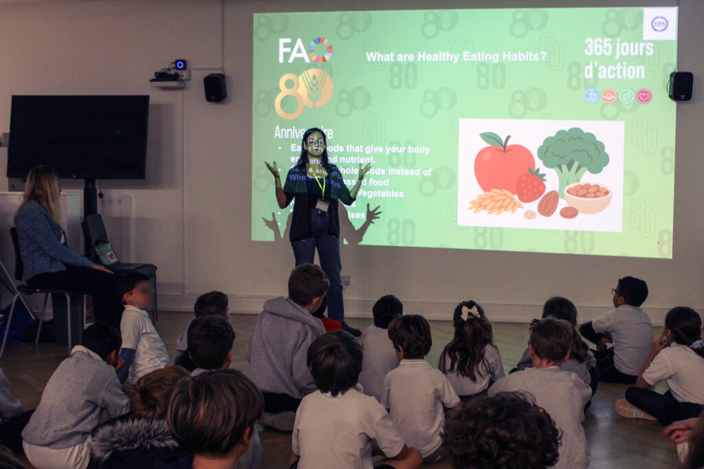 EIFA A woman stands in front of a projected screen, giving a presentation on healthy eating to a group of seated children. The screen shows a graphic of fruits, vegetables, and text about healthy habits.