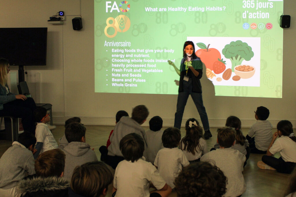 EIFA A woman gives a presentation about healthy eating habits to a group of children sitting on the floor, with a colorful slide showing fruits, vegetables, and grains projected on the wall behind her.