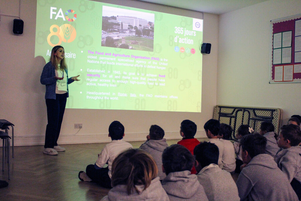 EIFA A woman stands in front of a projected presentation about the Food and Agriculture Organization, speaking to a group of seated children in a classroom. The slide highlights the FAO’s work and anniversary.