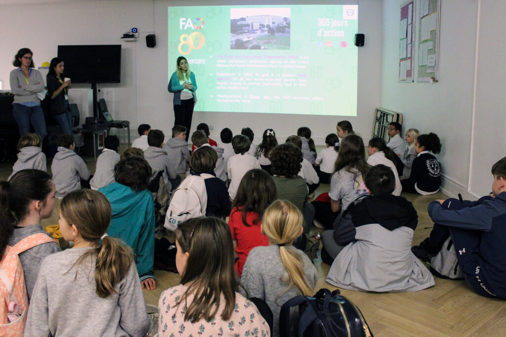 EIFA A group of children sit on the floor facing a screen as two women give a presentation in a brightly lit classroom. The screen displays colorful text and images about a project or event.